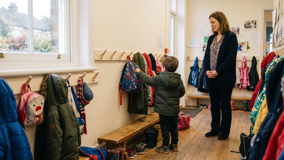 A young child in a coat reaches up to hang a small rucksack on a peg in a nursery cloakroom, seen from behind. A nursery worker stands a few steps back with her hands clasped, watching and letting the child manage it. The wall is lined with low pegs, coats and boots.